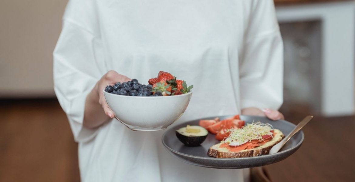 Person holding a bowl of fresh berries and a plate with avocado toast, displaying a healthy breakfast spread.
