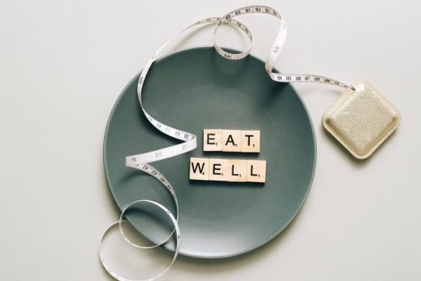 Flat lay of scrabble tiles spelling 'eat well' on a plate with a tape measure, promoting healthy eating.